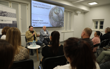 Dwaj mężczyźni siedzą na krzesłach przed ekranem; prelegent w beżowej kurtce przemawia do mikrofonu, gestykulując w stronę widowni