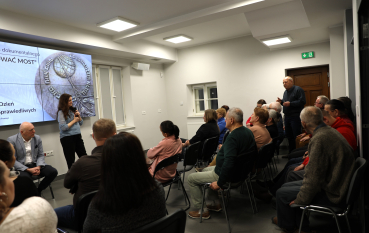 Kobieta z mikrofonem przemawia do siedzącej grupy; w tle jeden z uczestników stoi i zabiera głos w dyskusji.