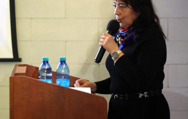 Zbliżenie na kobietę przemawiającą przy drewnianej mównicy; na blacie stoją dwie butelki wody.