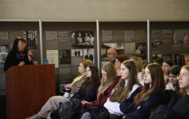 Widok z boku na prelegentkę przy mównicy i rzędy zasłuchanych uczniów na tle plansz edukacyjnych.