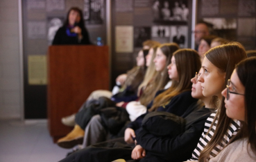 Uczniowie siedzący w rzędach, pokazani z profilu, patrzą w stronę ekranu z prezentacją, w tle nieostra sylwetka prelegentki.