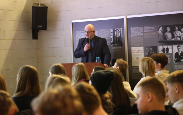 Mężczyzna w okularach i ciemnym garniturze przemawia do mikrofonu, stojąc za mównicą przed grupą słuchaczy.