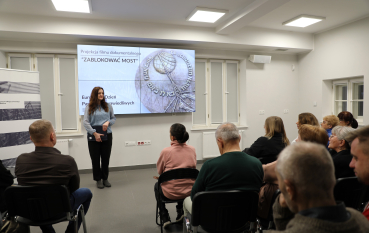 Kobieta stoi przed ekranem z napisem „Projekcja filmu dokumentalnego ZABLOKOWAĆ MOST” i przemawia do siedzącej publiczności.