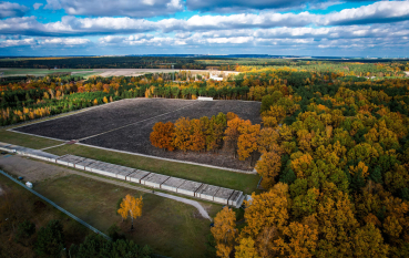 The aerial view reveals the museum’s geometric layout. The central area is dominated by a vast expanse of grey aggregate, which contrasts with the vibrant colours of the autumnal forest. A long, concrete museum building can be seen along one of the edges.
