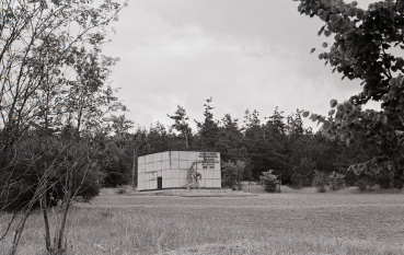 An archive black-and-white photograph of the first memorial to the victims of the Bełżec extermination camp. In a clearing surrounded by forest stands a concrete cuboid bearing a commemorative inscription and a sculpture depicting two emaciated figures clasped together. The scene is captured through the branches of the trees.