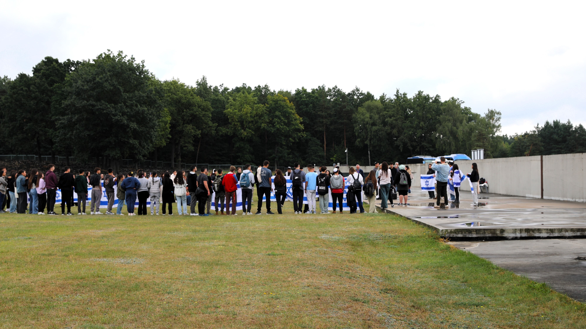 A wide shot showing a large group of young people gathered in the open grounds of the Museum and Memorial Site in Bełżec. The participants are standing in a large semicircle on a grassy area. They are holding outstretched blue and white Israeli flags bearing the Star of David. A wall of trees can be seen in the background