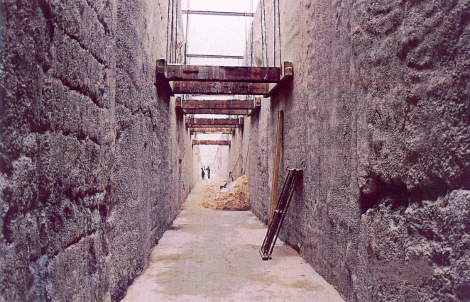 A colour photograph from December 2003 showing the construction of the ‘Szczelina’ in Bełżec. A view from inside a deep, narrow trench with bare concrete walls. Wooden struts are stretched across the passageway, and the silhouettes of workers can be seen in the distance.