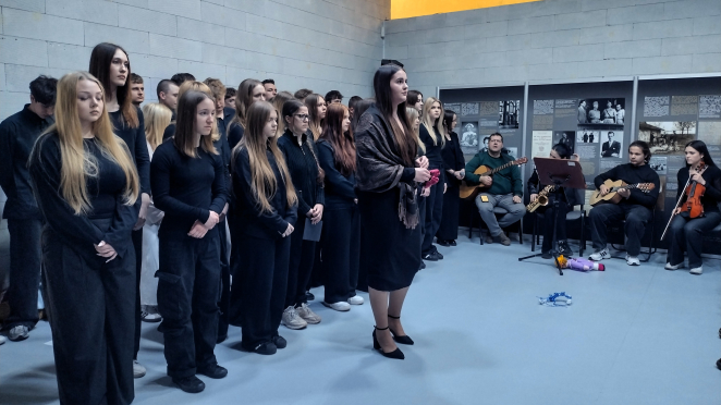 Grupa młodych ludzi ubranych na czarno stoi w surowym pomieszczeniu o betonowych ścianach. W tle widoczne są tablice edukacyjne ze zdjęciami i tekstem oraz zespół muzyczny z gitarzystami, saksofonistą i skrzypaczką. Na pierwszym planie wyróżnia się kobieta w ozdobnej chuście, a na podłodze leżą symboliczne przedmioty.