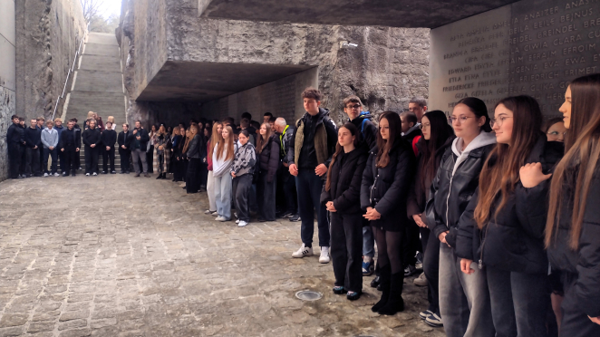 Młodzież stoi w skupieniu wewnątrz Niszy Ohel – przejmującej części upamiętnienia, stoją na tle ściany wyłożonej rzędami imion ofiar.