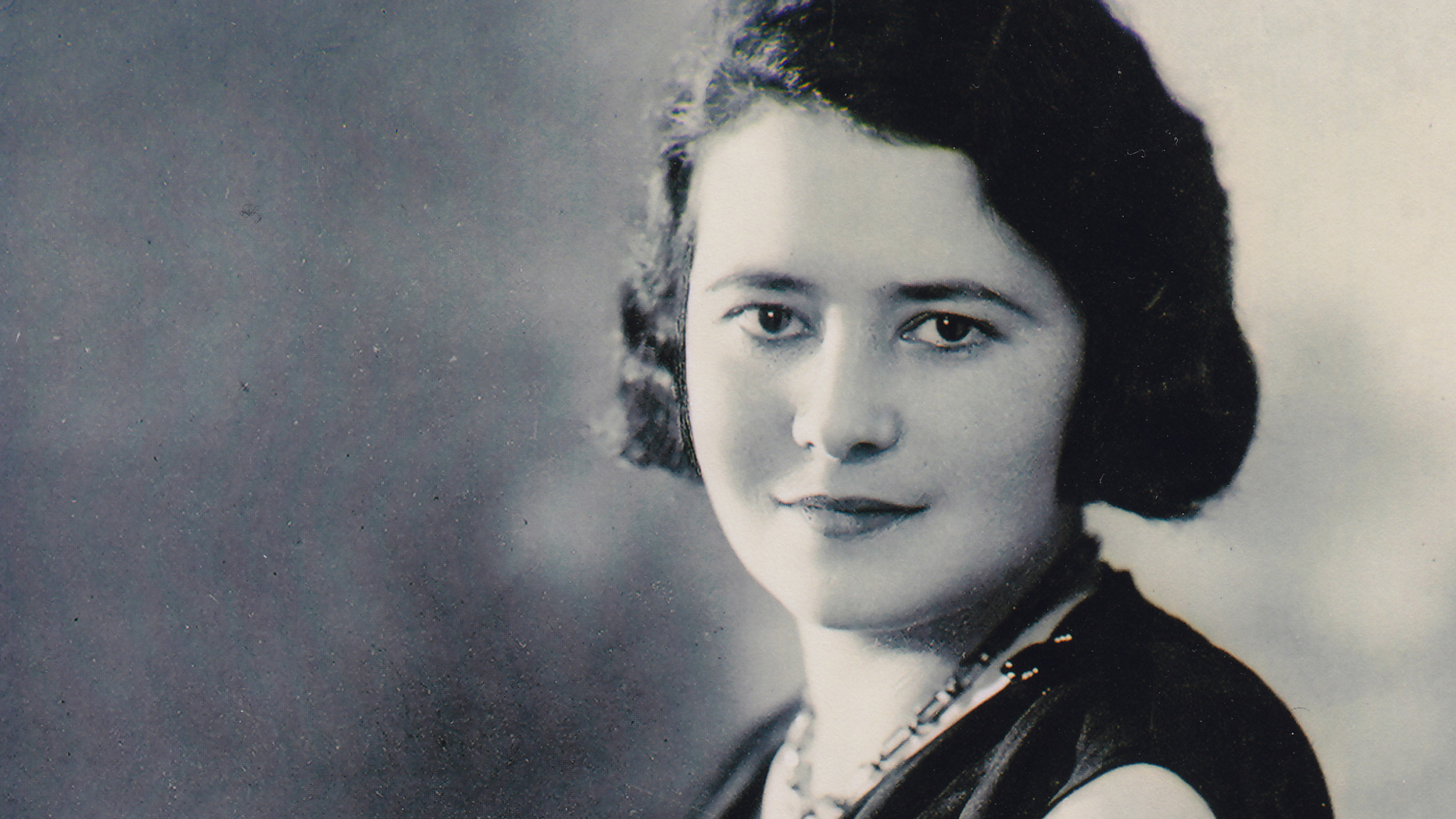 Czarno-biały, studyjny portret młodej kobiety o ciemnych, falowanych włosach sięgających do uszu. Kobieta siedzi bokiem, lekko uśmiechając się w stronę obiektywu; ma na sobie elegancką, ciemną suknię bez rękawów z drapowaniem przy dekolcie, naszyjnik oraz zegarek na lewym nadgarstku. Jej dłonie są splecione na kolanach.