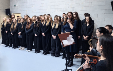 Kobieta w ciemnoniebieskiej sukience przemawia do mikrofonu, trzymając granatową teczkę. Za nią stoi grupa młodzieży w czarnych strojach.