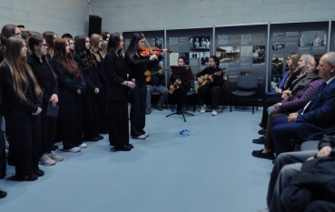 Szerokie ujęcie sali w Muzeum i Miejscu Pamięci w Bełżcu. Po lewej grupa młodzieży, w głębi zespół muzyczny, a po prawej siedzący goście. Na środku stoją skrzypaczka grająca na skrzypcach oraz młoda dziewczyna z mikrofonem. Na ścianach ekspozycja historyczna.