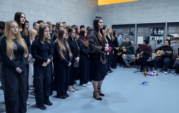 Grupa młodzieży, przed nią stoi dziewczyna w ozdobnej chuście, w tle skrzypaczka i gitarzyści. Na podłodze leżą symboliczne rekwizyty – niebieski wianek i lalka.