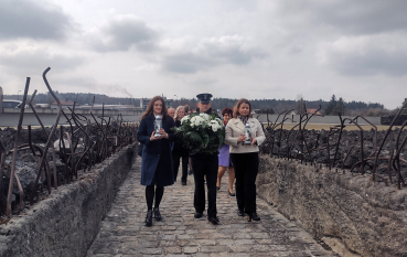 rzy osoby – dwie kobiety w płaszczach i mężczyzna w mundurze – idą brukowaną ścieżką (Szczeliną-Drogą), niosąc biały wieniec i znicze