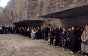 Duża grupa uczestników stoi w milczeniu w monumentalnej przestrzeni Niszy Ohel. Wokół surowe betonowe ściany.