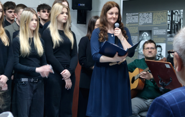 Zbliżenie na kobietę w ciemnoniebieskiej sukience. Przemawia do mikrofonu, trzymając granatową teczkę. Za nią stoi grupa młodzieży w czarnych strojach