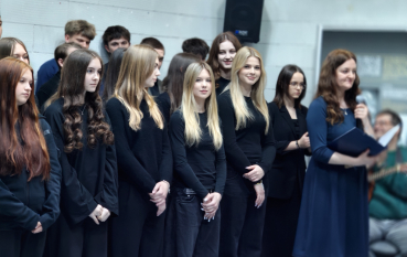 Grupa młodych osób stoi w skupieniu wzdłuż jasnej, betonowej ściany. Większość ubrana jest w jednolite, czarne swetry lub bluzy. Po prawej stronie dorosła kobieta w ciemnobłękitnej sukience przemawia do mikrofonu, trzymając w dłoniach granatową teczkę z tekstem