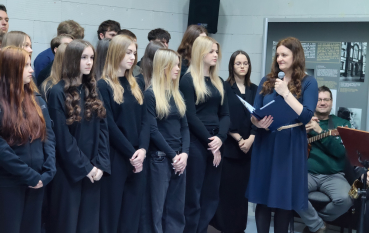 Grupa młodych osób stoi w skupieniu wzdłuż jasnej, betonowej ściany. Większość ubrana jest w jednolite, czarne swetry lub bluzy. Po prawej stronie dorosła kobieta w ciemnobłękitnej sukience przemawia do mikrofonu, trzymając w dłoniach granatową teczkę z tekstem