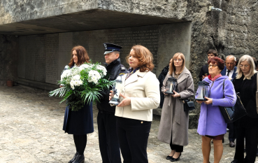 Grupa osób stoi w Nichy Ohel za nimi monumentalna, surowa ściana z betonu, na której wyryto liczne imiona. Na pierwszym planie dwie kobiety i mężczyzna w mundurze trzymają białe wieńce oraz znicze w ozdobnych lampionach.