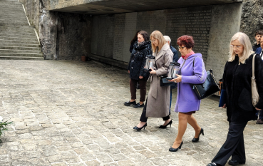 Uczestnicy uroczystości składają lampiony w tle betonowa tablica z imionami. Na pierwszym planie cztery kobiety, dwie niosą zapalone znicze w czarnych lampionach. W tle widoczne są betonowe schody prowadzące w górę.