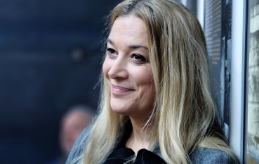 Portret uśmiechniętej kobiety o długich blond włosach. Kobieta ma na sobie szarą marynarkę z czarnym elementem ozdobnym pod szyją. W rękach trzyma białe dokumenty oraz telefon w jasnoróżowym etui. W tle widoczny jest fragment wystawy oraz rozmyte sylwetki innych osób.