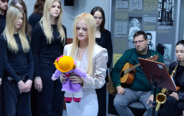 Scena symboliczna. Młoda kobieta w białym stroju stoi na środku sali, trzymając w dłoniach fioletową, włóczkową lalkę w żółtym kapeluszu. U jej stóp leży wiklinowy kosz wypełniony białymi i niebieskimi kwiatami. W tle siedzi mężczyzna z gitarą, a obok widoczna jest saksofonistka..