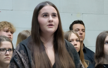 Młoda kobieta o długich, brązowych włosach z narzuconą na ramiona ozdobną, ciemną chustą z frędzlami. Trzyma w dłoniach różowe maleńkie buty dziecięce wykonane z materiału, patrząc w górę z wyrazem głębokiego przejęcia. W tle grupa młodych ludzi.