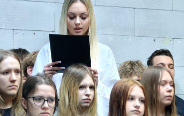W górnej części kadru blondynka w białym stroju czyta z czarnej karty. Poniżej widać twarze młodych kobiet o poważnych, zadumanych wyrazach twarzy, patrzących przed siebie. Jedna z nich ma długi warkocz z różową gumką.