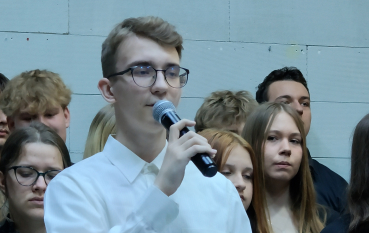 Portretowe ujęcie młodego mężczyzny w okularach i eleganckiej białej koszuli, który przemawia do mikrofonu. W tle widać twarze innych uczestników.