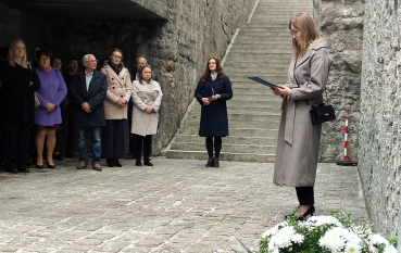 obieta w długim beżowym płaszczu stoi bokiem do kamiennej ściany i czyta z dokumentu trzymanego w granatowej teczce. U jej stóp leży duży wieniec z białych kwiatów oraz zapalony znicz. W głębi, pod betonowym zadaszeniem, stoi grupa osób obserwująca ceremonię. W tle widać wysokie, surowe ściany i schody.