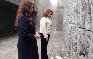 Dwie kobiety w płaszczach (granatowym i jasnym) stoją w głębokim skłonie przed kamiennym murem, oddając hołd. Przed nimi, na bruku, leży wieniec z białych kwiatów oraz płonący znicz.