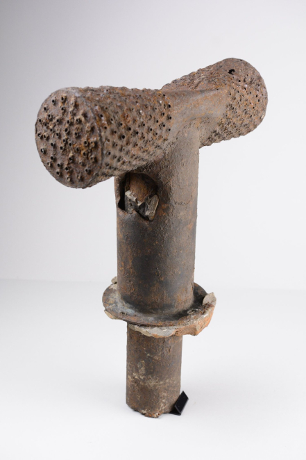 Silnie skorodowany, metalowy fragment instalacji w kształcie litery „T”, odnaleziony na terenie byłego niemieckiego obozu zagłady. Artefakt składa się z pionowej rury z szerokim kołnierzem u dołu oraz poprzecznej głowicy z licznymi, małymi otworami na końcach, przypominającymi sitko. Obiekt jest pokryty grubą warstwą rdzy i wżerami, a w górnej części trzonu widoczne jest pęknięcie.