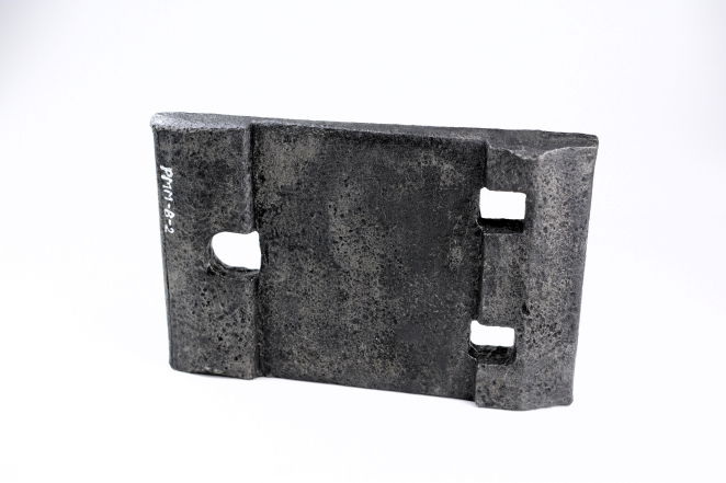 Metalowy podkład kolejki wąskotorowej, ustawiony pionowo na jasnym, jednolitym tle. Przedmiot ma formę ciężkiej, prostokątnej płyty wykonanej z ciemnego, chropowatego żeliwa lub stali z widocznymi śladami korozji i wżerów. Na bocznej krawędzi widnieje biały napis inwentarzowy „PMM-B-2”. Konstrukcja podkładu zawiera charakterystyczne wyżłobienia i otwory montażowe: po lewej stronie znajduje się jeden większy, owalny otwór, natomiast po prawej dwa mniejsze, prostokątne wycięcia.