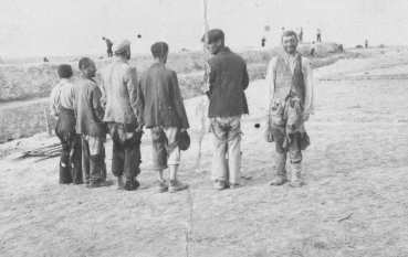 Czarno-biała fotografia przedstawia grupę sześciu mężczyzn - Żydów. Pięciu stoi tyłem do obiektywu jeden przodem na piaszczystym, otwartym terenie. Mężczyźni są ubrani w bardzo zniszczone marynarki oraz spodnie. Stoją obok głębokiego wykopu, którego krawędzie są wzmocnione drewnianymi szalunkami i deskami. Wykop to rów przeciwczołgowy.