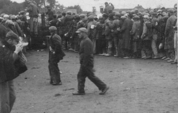 Duża grupa ludzi na rozległym, płaskim placu. Po prawej stronie formuje się długa kolejka osób stojących jedna za drugą, ciągnąca się w głąb kadru. Wiele osób ma na sobie nakrycia głowy i niesie bagaż podręczny lub chlebaki. Po lewej stronie widać mniejsze skupiska ludzi oraz coś, co przypomina drewniane wozy lub prowizoryczne punkty wydawania posiłków.