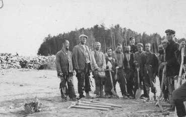 Czarno-biała fotografia portretowa grupy dziesięciu mężczyzn pozujących do zdjęcia w lesie. Mężczyźni stoją w jednym rzędzie, patrząc w stronę aparatu. Są ubrani w cywilne, znoszone ubrania: marynarki, płaszcze i czapki. Wielu z nich trzyma w dłoniach narzędzia pracy – łopaty o długich trzonkach oraz siekierę. Przed nimi, na ziemi pokrytej igliwiem, leży więcej narzędzi. Mężczyźni mają zmęczone twarze. Za ich plecami gęsto rosną młode sosny.