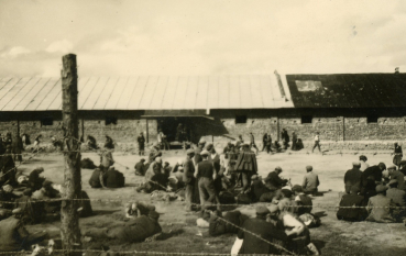 Czarno-białe zdjęcie przedstawiające grupę ludzi siedzących i leżących na ziemi na ogrodzonym terenie z drutem kolczastym; w tle długi, niski budynek z cegły.