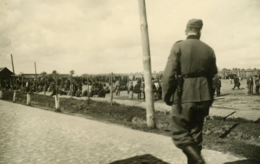 Czarno-białe zdjęcie przedstawiające umundurowanego strażnika idącego wzdłuż ogrodzenia, za którym znajduje się duża grupa ludzi zgromadzonych na otwartym terenie.