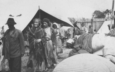 Czarno-białe zdjęcie dokumentalne przedstawiające grupę dorosłych i dzieci na otwartej przestrzeni otoczonej wysokim, drewnianym płotem. Na pierwszym planie widać kobietę w kraciastej chuście narzuconej na ramiona oraz mężczyznę w kapeluszu i ciemnej kurtce. W głębi widoczne są liczne namioty, prowizoryczne szałasy oraz duże białe worki lub tobołki z rzeczami osobistymi.