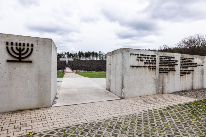 Betonowe obramowanie z metalową inskrypkcją, w tle pomnik Nisza Ohel 