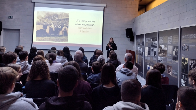 Widok z tyłu sali na publiczność i prelegentkę w czerni. Na ekranie wyświetlany jest slajd o rodzinie Ulmów z archiwalnym zdjęciem.