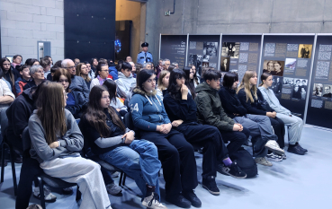 Grupa młodzieży siedząca w rzędach, słuchająca wykładu. W tle widoczne betonowe ściany muzeum i fragmenty wystawy z fotografiami.