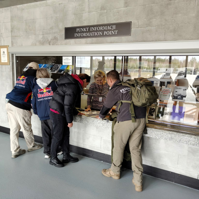 Czworo turystów z plecakami (dwaj w kurtkach Red Bull) stoi przy okienku punktu informacyjnego, rozmawiając z pracownicą.