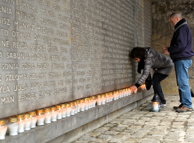 Kobieta pochyla się, zapalając znicz przy długim rzędzie płonących lampionów ustawionych wzdłuż betonowej ściany z wyrytymi imionami, obok stoi mężczyzna.