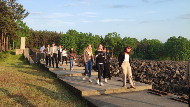 Grupa młodych ludzi schodzi po betonowych schodach, mijając rozległe pole ciemnych, ostrych kamieni. Po lewej stronie widać las, a w oddali betonową ścianę.
