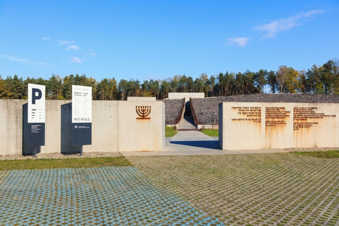 Widok z przodu na wejście do muzeum, z betonowymi ścianami, tablicami informacyjnymi, symbolem menory.