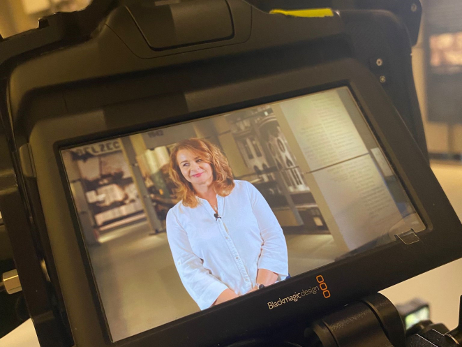 Widok zza kulis na kamerę filmową Blackmagic Design, na której ekranie widać nagrywaną kobietę z rudymi włosami w białej koszuli, uśmiechającą się.