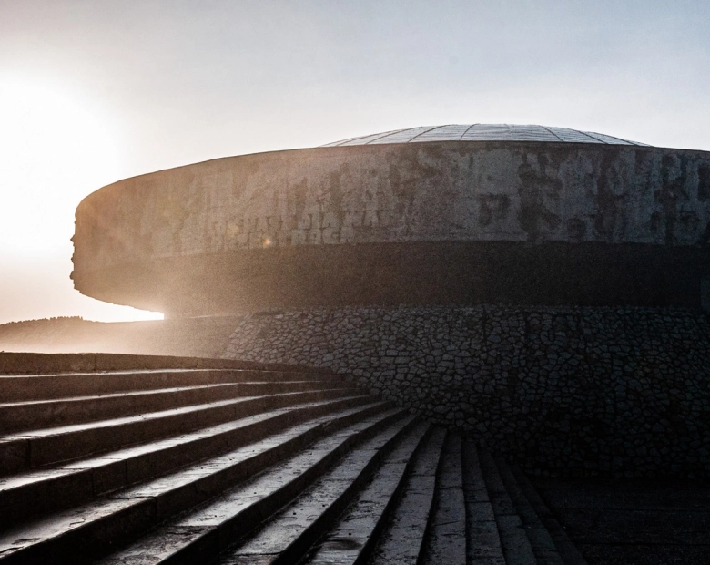 Kopuła Mauzoleum w muzeum na Majdanku widziana od dołu, oświetlony niskim światłem słońca. Na pierwszym planie prowadzą do niego szerokie kamienne schody, a sylwetka budynku odcina się na tle jasnego nieba.