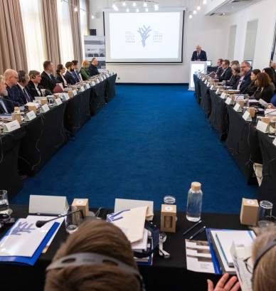 Ludzie elegancko ubrani w sali konferencyjnej siedzący przy stołach ułożonych w podkowę, jedna osoba przy mównicy, na ścianie wyświetlany baner.   