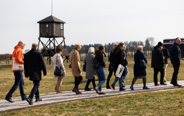 Grupa osób idzie przez teren byłego obozu koncentracyjnego na Majdanku, w tle wieża wartownicza. 